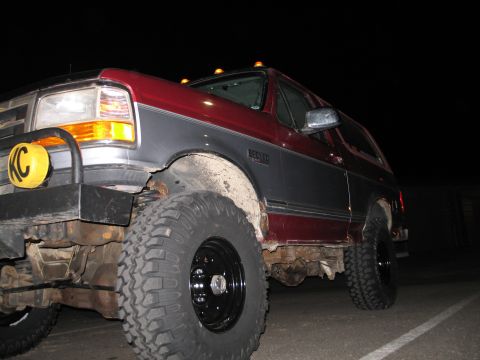 1995 Ford Bronco - A lifted two-tone maroon and gray Ford Bronco SUV photographed at night from a low front-3/4 angle, showing large mud-terrain tires on blac…