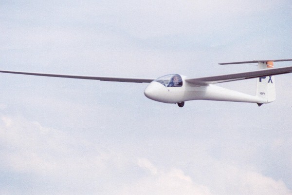 White single-seat glider in flight with FX marking on the tail against a cloudy sky