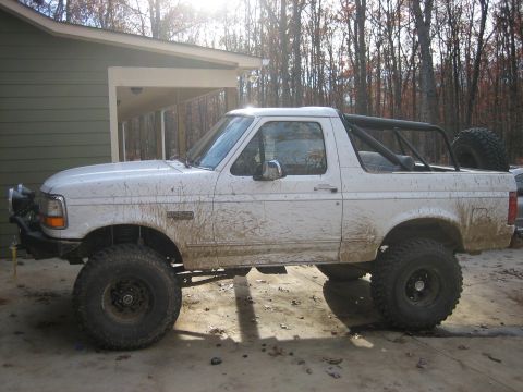 1996 Ford Bronco - A muddy 1996 Ford Bronco with a roll cage and large off-road tires is parked outside a building.