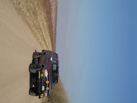 1995 Ford Bronco - A dark maroon/burgundy Ford Bronco SUV parked on a dirt road in an open prairie setting, shown from the front-quarter angle with a brush gu…