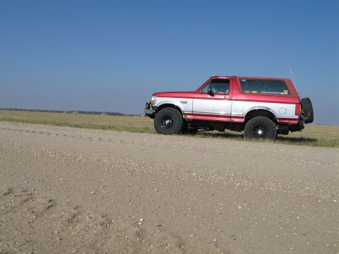 1995 Ford Bronco - A red and white two-door Ford Bronco full-size SUV with a lift kit and off-road tires on black wheels, parked in profile on a gravel road b…