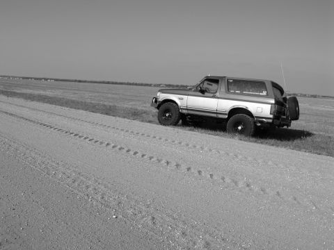 1995 Ford Bronco - Black-and-white side view of a two-tone full-size Ford Bronco with a hardtop parked in a flat grass field beside a gravel road. It's lifted…
