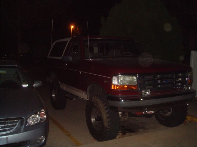 Lifted burgundy Ford Bronco with white top parked at night beside a sedan