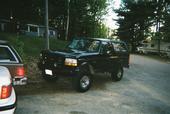 1995 Ford Bronco - A black 1995 Ford Bronco is parked on a dirt road, with trees and houses in the background.