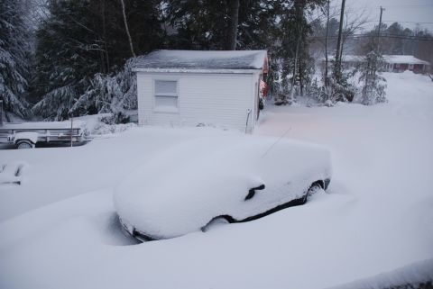 1996 Ford Bronco - A vehicle is completely covered in snow, with a small white shed and trees in the background.