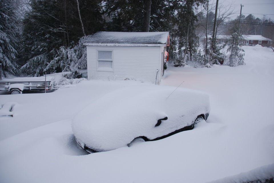 Car buried in snow next to a white shed. Trailer visible to the left