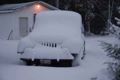 1996 Ford Bronco - A vehicle is completely covered in snow, parked in front of a house during a winter storm.