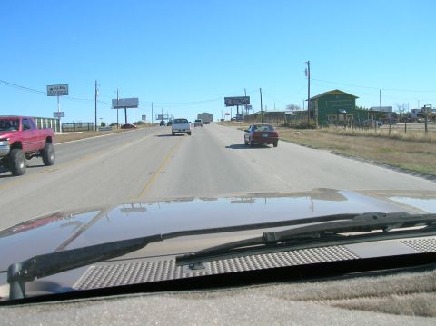 1996 Ford Bronco - A lifted red pickup truck drives down a highway on a clear day, with other vehicles and billboards visible.