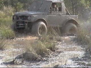 1987 Suzuki Samurai - This off-road vehicle is covered in mud as it drives through a watery path surrounded by vegetation.