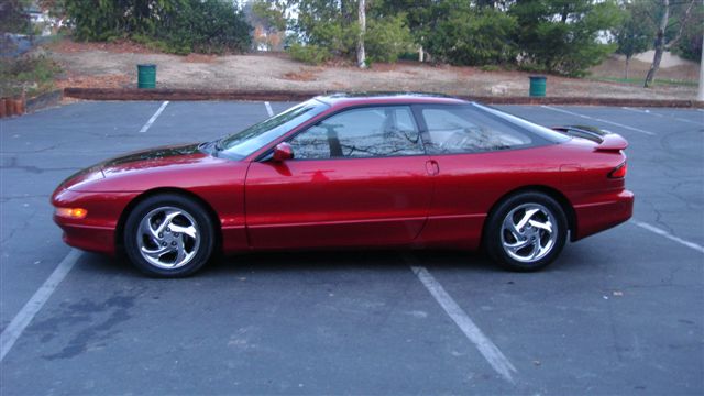 Red coupe parked in a lot, side view. Chrome wheels, spoiler, tinted windows