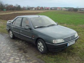 1989 Ford Bronco - A dark grey Peugeot 405 sedan is seen from a three-quarter front view, parked on a dirt road with fields in the background.