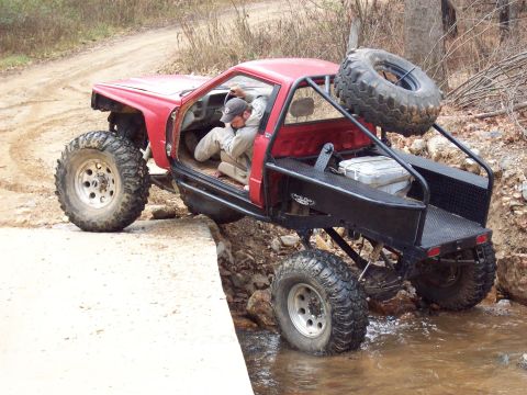 1978 Ford Bronco - Modified red off-road vehicle with oversized tires and a custom roll cage is driven through a shallow stream.