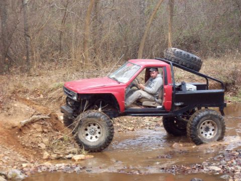 1978 Ford Bronco - A man drives a modified red pickup truck through a muddy creek bed surrounded by trees.