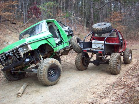 1978 Ford Bronco - Two heavily modified off-road trucks are on a dirt trail in a wooded area, with one appearing to be stuck.