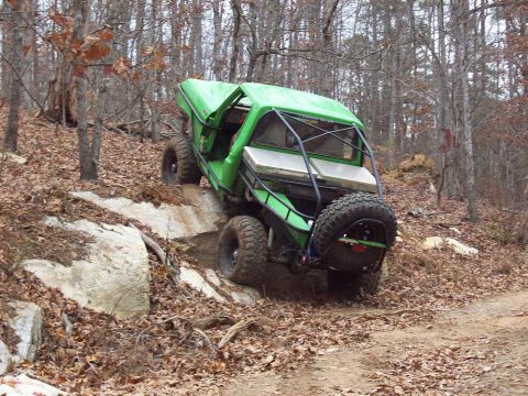 1978 Ford Bronco - A modified green off-road vehicle navigates a challenging rocky terrain on a wooded trail.