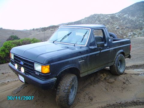 1990 Ford Bronco - A black Ford Bronco with off-road tires navigates a muddy terrain under an overcast sky.
