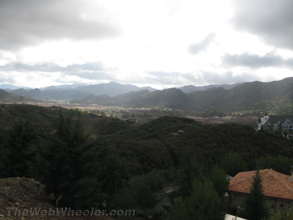 Overcast sky above rolling hills and distant mountains, with houses on the right and trees in the foreground