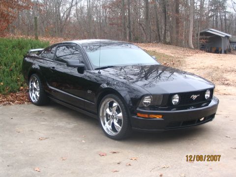 2007 Ford Mustang - A black Ford Mustang GT is parked on a concrete driveway with a wooded area behind it.