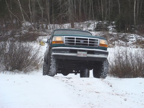 1996 Ford Bronco - A lifted green Ford truck navigates a snowy, wooded area with its front end angled towards the camera.