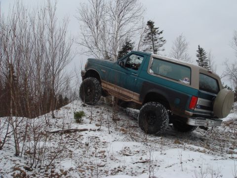 1996 Ford Bronco - Teal Ford Bronco with tan top and large tires climbs a snowy hill.