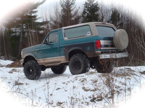 1996 Ford Bronco - A teal and tan 1996 Ford Bronco with oversized tires navigates a snowy, wooded landscape.