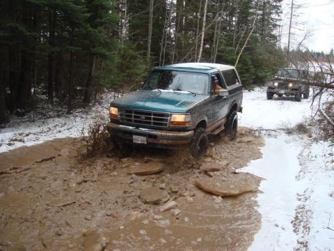 1996 Ford Bronco - Two off-road vehicles navigate a challenging muddy and snowy path in a wooded area.