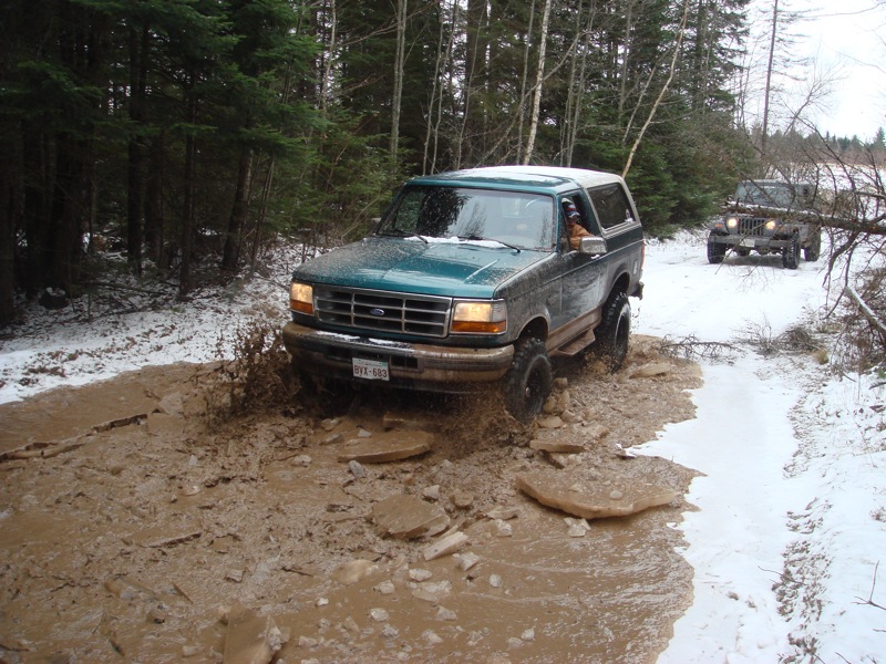 1996 Ford Bronco driving through a muddy trail with snow on the ground; another Jeep follows behind