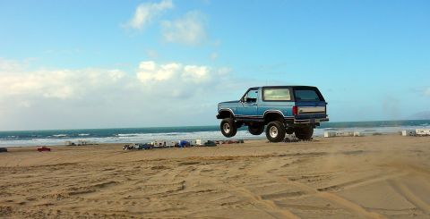 1984 Ford Bronco - A blue Ford Bronco is captured mid-air on a beach, with the ocean and several trailers visible in the distance.