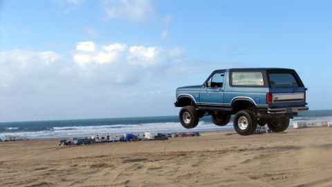1984 Ford Bronco - Blue 1984 Ford Bronco airborne over a beach with RVs parked nearby.