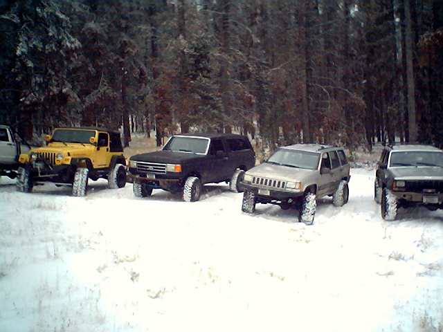 Several off-road vehicles parked in a snowy forest. Includes a yellow Jeep, a black SUV, and two silver/gray SUVs
