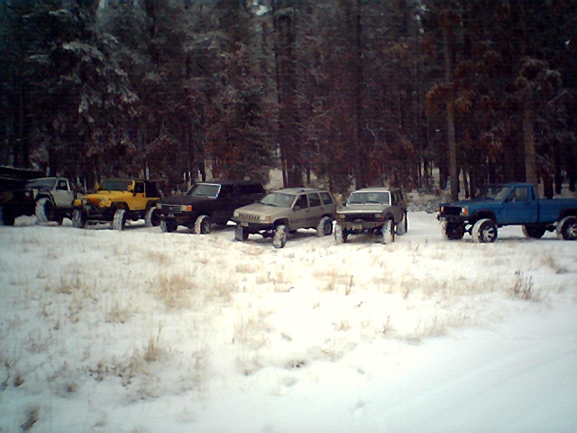 Several modified SUVs and a blue pickup truck parked in a snowy forest clearing
