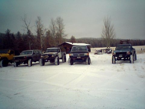 1990 Ford Bronco - A group of off-road vehicles are parked in a snowy landscape near a rustic cabin.