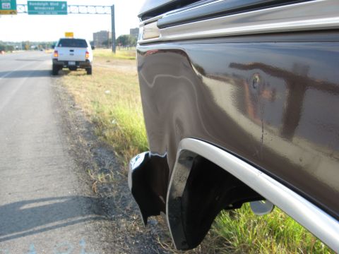 1979 Ford Bronco - A close-up of a dark brown vehicle's rear quarter panel, showing chrome trim and a damaged bumper. Another vehicle is visible on the road a…