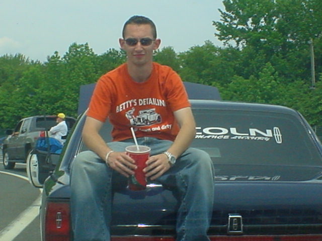 Man in orange shirt sits on trunk of dark car, holding a red cup. Another car and person visible in background