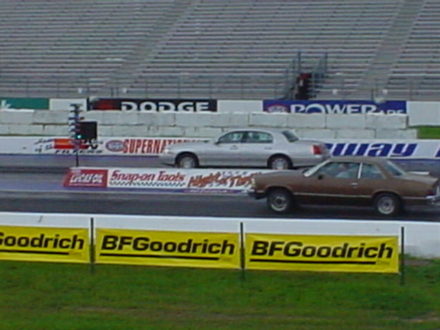 Two cars on a drag strip, one silver sedan and one brown coupe, with grandstands and sponsor banners in the background