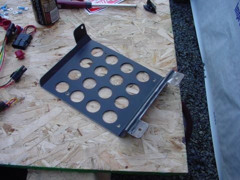 1979 Ford Bronco - A black metal battery tray with a pattern of circular holes is shown on a piece of oriented strand board.
