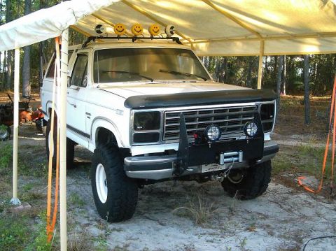 1986 Ford Bronco - White lifted Ford Bronco parked on sandy ground under a white canopy tent, viewed from the front three-quarter with a black hood bra, a roo…
