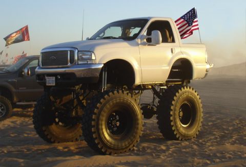 1970 Ford Ranchero - A heavily modified white pickup truck with massive tires and an American flag is parked on sandy terrain.