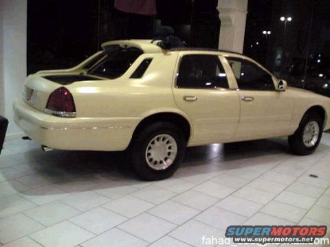 1972 Ford Ranchero - Cream/pale-yellow Ford Crown Victoria four-door sedan photographed from the 3/4 rear on white tile flooring inside a lit showroom at night.…