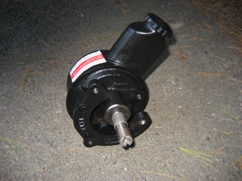 1991 Ford Bronco - A black power steering pump is shown resting on a paved surface with some pine needles scattered around.