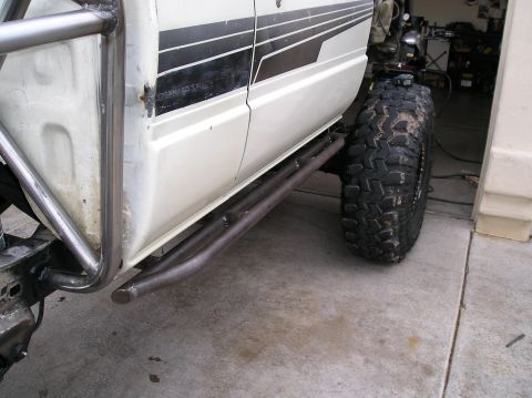 1986 Toyota Hilux - Low side view of a white pickup in a garage showing bare-steel tube rock sliders and a bent tube rear fender/exocage bar welded to the body…