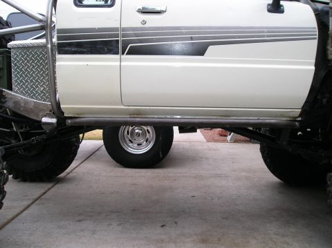 1986 Toyota Hilux - Ground-level side view of a lifted white pickup on a driveway, framed under the body to show a bare-steel tube rock slider, diamond-plate f…