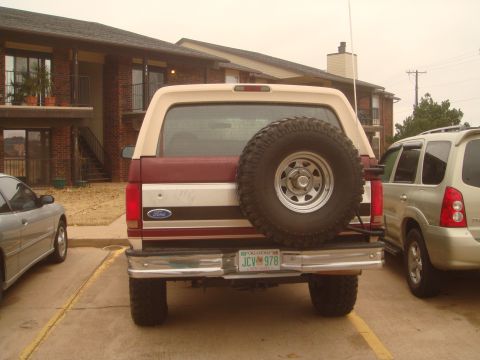1992 Ford Bronco - Straight-on rear view of a lifted burgundy Ford Bronco with a tan removable hardtop, parked between other vehicles at a brick apartment com…