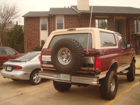 1992 Ford Bronco - Passenger-side rear three-quarter view of a lifted burgundy Ford Bronco with a tan removable hardtop, parked outside a brick apartment buil…