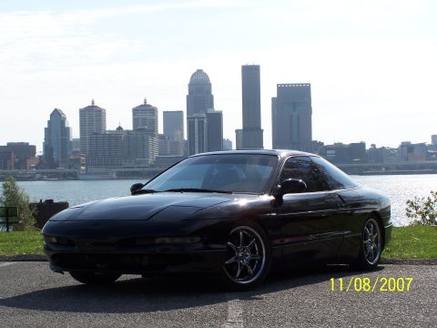 1995 Ford Probe - A black 1995 Ford Probe is parked near a waterfront, with a city skyline in the background.