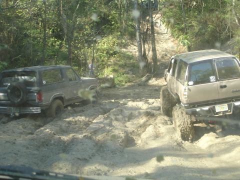 1989 Ford Bronco - Off-road vehicles struggling through a muddy terrain on a wooded trail.