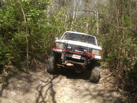 1989 Ford Bronco - White off-road SUV navigates a wooded trail with large tires and a modified front bumper.