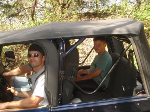 1989 Ford Bronco - Two people are inside a dark blue Jeep with a soft top, driving on a wooded trail.