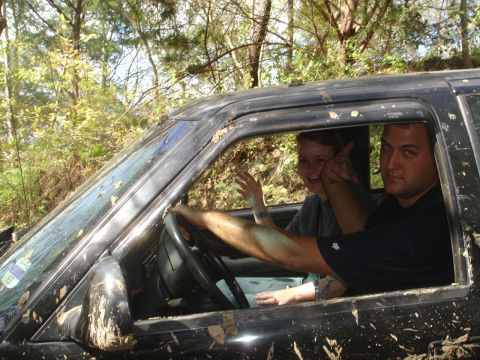 1989 Ford Bronco - Two people are inside a dirty vehicle, possibly off-roading, with trees visible through the windows.