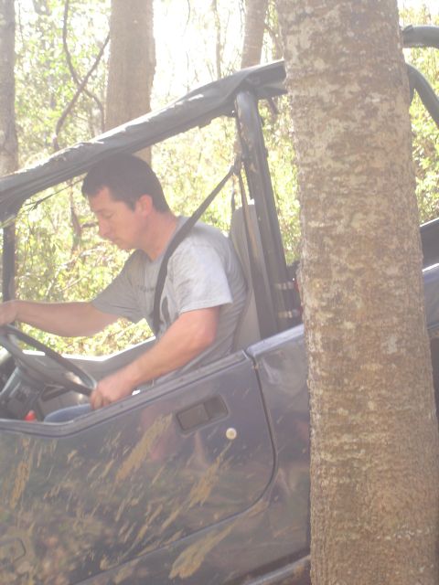 1989 Ford Bronco - Man drives a muddy off-road vehicle off-road, navigating around a tree.